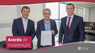 Na foto, estão o presidente do TCE/SC, conselheiro Adircélio de Moraes Ferreira Júnior (à direita), o prefeito de Blumenau, Mário Hidelbrandt (centro), que segura o acordo de cooperação técnica, e o conselheiro José Nei Ascari (à esquerda). Todos estão em pé, vestem paletó escuro e camisa clara. À frente da imagem, um retângulo bordô com o texto Acordo de cooperação, em fonte branca.