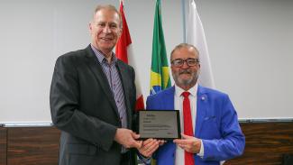 Foto do presidente do TCE/SC, Herneus De Nadal, e do presidente do TJSC, Altamiro de Oliveira. Ambos vestem terno. Eles estão em pé, um ao lado do outro, na sede do TCE/SC. Estão sorrindo para a foto enquanto seguram e mostram a menção honrosa recebida, uma placa de metal. 