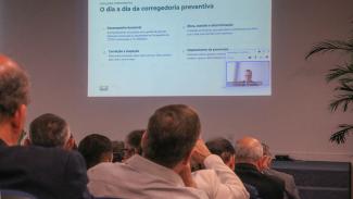 Banner horizontal com foto, em primeiro plano, da plateia assistindo à videoconferência com o conselheiro Adircélio, exibida no telão ao fundo. 