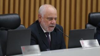 Foto do relator das contas do Governo do exercício de 2024, conselheiro Luiz Eduardo Cherem, durante sessão do Pleno. Ele é um homem branco, de cabelos e barba grisalhos. Está falando ao microfone, sentado em poltrona diante da bancada circular de autoridades que compõem o Plenário.