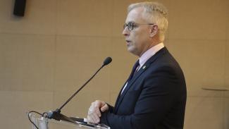 Banner horizontal com foto de perfil do diretor-geral de Controle Externo, Sidney Tavares, falando ao microfone durante sessão plenária. É um homem branco, de cabelos grisalhos. Veste terno e gravata e usa óculos de grau.