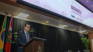 Foto do conselheiro Adircélio de Moraes Ferreira Júnior discursando no púlpito, no canto esquerdo do palco do auditório do TCE/SC. Ele é um homem branco, de cabelos curtos e castanhos, e usa óculos. Está vestindo um terno azul-escuro, camisa azul-claro e gravata roxa. Atrás dele, no lado esquerdo, as bandeiras do Brasil e de Santa Catarina. Acima, o painel eletrônico, onde aparece a logo do evento. 