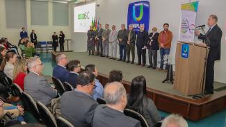 Foto da cerimônia de adesão do município de Lages ao Acerta SC. Em primeiro plano, de costas, há participantes do evento que estão sentados em poltronas do auditório da Uniplac. À sua frente, no palco, há 10 autoridades, em pé, uma ao lado da outra. Entre os presentes, o conselheiro Aderson Flores e o prefeito de Lages, Antônio Ceron. Falando ao púlpito, está o presidente do TCE/SC, Herneus De Nadal.  