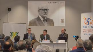 Banner horizontal com foto de três homens sentados diante de uma bancada, no palco. Entre eles, o conselheiro Adircélio Ferreira Júnior e o professor Antonio Arias Rodríguez. À frente deles, o público que assiste ao painel, de costas. Ao fundo, atrás dos painelistas, no telão, foto, em preto e branco, do conselheiro Salomão Ribas Junior. 