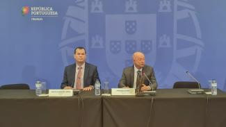 Foto da Mesa do Seminário, onde aparecem sentados o conselheiro Adircélio de Moraes Ferreira Júnior (TCE/SC) e o conselheiro Fernando Toledo (TCE/AL), este último falando ao microfone. A parede de fundo é na cor azul e contém uma grande imagem do Brasão de Portugal em tom mais claro. Ainda na parede, no canto superior esquerdo aparece pequena bandeira de Portugal e a expressão “República Portuguesa – Finanças”