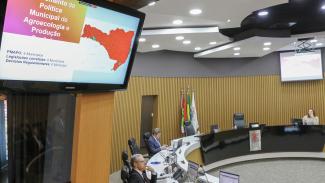 Foto da sessão plenária. Em primeiro plano, uma televisão projeta a apresentação do novo painel de agroecologia do Farol TCE/SC. Em segundo plano, os conselheiros sentados diante da bancada circular do Pleno. Na foto, o conselheiro substituto Gerson dos Santos Sicca e o conselheiro José Nei Ascari.