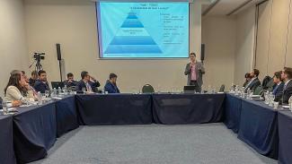 Foto do servidor do TCE/C Alessandro Marinho está fazendo a apresentaação sobre o VigIA.  As cadeiras e mesas estão dipostas em formato de U. Ao fundo do palestrante, um telão exibe a apresentação dele.