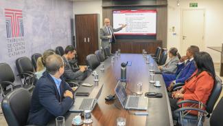 Foto da sala onde ocorreu a reunião. Ao fundo, o diretor Sidney Tavares faz a apresentação diante de um monitor de televisão. À frente, a mesa de reuniões onde estão sentados os demais diretores do TCE/SC, de um lado, e os integrantes do TC de Angola, do outro. 