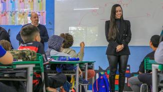 Banner horizontal com foto de uma sala de aula com alunos sentados em suas carteiras durante uma atividade escolar. Em destaque, a auditora Letícia Spindola de Faria está em pé à frente da turma, sorrindo e interagindo com os estudantes. Ao fundo, próximo ao quadro branco, o auditor Renato Bossle Miguel acompanha a atividade. 