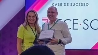 Foto do assessor de Governança Estratégica de Tecnologia da Informação do TCE/SC, Jairo Wensing, recebendo o prêmio Case de Sucesso em evento. Ele está ao lado de uma mulher, segurando o a premiação, mostrando-a para a foto. Ambos sorriem. Ao fundo, projeção do texto “Case de sucesso” e o logo do TCE/SC.