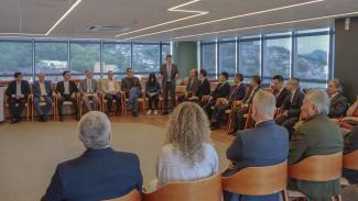 Foto horizontal do vice-presidente José Nei Ascari, em pé, ao centro da sala, discursando e gesticulando para os demais participantes, que estão sentados em cadeiras dispostas em formato semicircular. Entre os presentes estão autoridades do Tribunal e representantes da instituição chinesa, em sua maioria vestindo trajes sociais. O ambiente é amplo e moderno, com piso de madeira, iluminação linear no teto e grandes janelas de vidro que permitem visualizar uma paisagem arborizada e construções ao fundo.