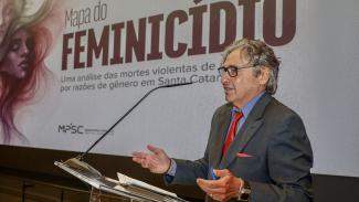 Banner horizontal com foto do conselheiro Aderson Flores discursando em um púlpito, com microfone, durante o evento. Ele veste terno escuro com gravata vermelha, usa óculos de grau e gesticula com as mãos enquanto fala. Ao fundo, há um telão com o título “Mapa do Feminicídio”