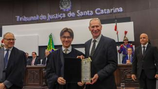 Foto horizontal do presidente do TCE/SC, conselheiro Herneus De Nadal, recebendo a homenagem durante sessão solene. Ele está ao lado de um integrante do TRE-SC. Ao fundo, na parede, o letreiro do Tribunal de Justiça de Santa Catarina. 