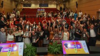Foto horizontal que mostra os formandos do curso do EJA do TCE/SC em meio à plateia, no auditório do TCE/SC, erguendo os certificados recebidos. Há várias pessoas em pé, em frente às cadeiras vermelhas.  