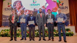 Banner horizontal com foto dos participantes do evento. Há seis homens, em pé, um ao lado do outro, em frente ao banner com logo do primeiro Enapi. Entre os presentes, o fotógrafo Guto Kuerten e os auditores fiscais de controle externo Edelvan da Conceição e Rafael Scherb. Eles seguram certificados.    