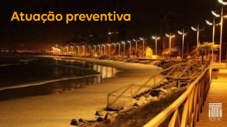 Foto noturna da praia de Navegantes, com a avenida principal à direita, iluminada por seguidos postes, que dão à foto uma coloração dourada. A esquerda, a faixa de areia e a água do mar, que reflete as luzes. No canto superior esquerdo, sob o céu escuro, o título “Atuação preventiva”, em fonte alaranjada. No canto inferior direito, a logo do TCE, na cor branca. 