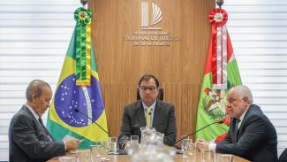 Imagem que mostra três autoridades sentadas diante de uma mesa em reunião. À esquerda, o governador Jorginho Mello, à direita, o conselheiro do TCE/SC Luiz Eduardo Cherem, e, ao centro, o desembargador Francisco Oliveira Neto. Atrás deles, na parede, logo do Tribunal de Justiça de Santa Catarina e as bandeiras do Brasil e de Santa Catarina. 