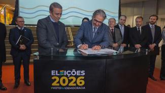 Banner horizontal com foto do conselheiro Aderson Flores assinando o documento, acompanhado por outros representantes ao redor da mesa. Ao fundo, outras autoridades observam a cerimônia. Na parte frontal do púlpito, há a inscrição "Eleições 2026".