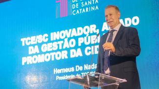 Foto do presidente do TCE/SC, conselheiro  Herneus de Nadal, diante do púlpito de acrílico. Ele é um homem branco, está vestindo terno azul-marinho, camisa rosa e gravata azul. Ao fundo, imagem de telão com a logo do TCE e o tema da palestra. 