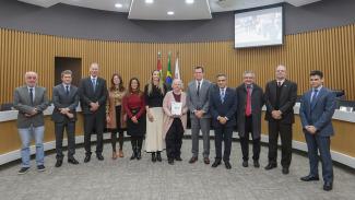 Banner horizontal com foto de 12 pessoas em pé, no Plenário do TCE/SC, durante entrega dos exemplares a Hanne Kristoffersen. Entre os presentes, além da bibliotecária, o presidente, o vice-presidente e o corregedor-geral do TCE/SC, conselheiros Herneus De Nadal, José Nei Ascari e Adircélio Ferreira Júnior, o presidente da Atricon, Edilson Silva, e a integrante da equipe executiva da revista, Juliana Fritzen. 