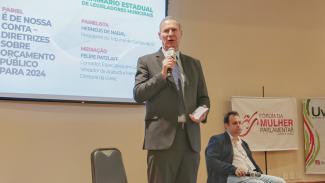 Foto do Presidente do TCE/SC, Herneus De Nadal, de pé, palestrando no auditório da Assembleia Legislativa. Ele é um homem branco, está de terno cinza claro, camisa clara e gravata azul-marinho. Ele segura o microfone com a mão direita. Ao fundo aparece parte do telão projetando informações sobre o Seminário, e ainda dois banners do evento. Ao lado, aparece sentado o vereador de Arabutã e membro da diretoria da UVESC, Felipe Patzlaff. 