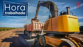 Foto de uma estrada de terra batida, onde estão um caminhão-caçamba, à frente, e uma retroescavadeira, atrás, colocando aterro na caçamba. No canto superior esquerdo, uma tarja azul com o texto “Hora trabalhada”. E no canto superior direito, a logo do TCE/SC. 