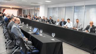 Banner com foto da reunião que contou com a participação de diversos conselheiros de tribunais de contas do Brasil. Eles estão sentado em volta de mesas, dispostas no formato de U. Sobre as mesas, há taças, notebooks e papéis.