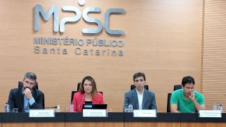 Descrição da imagem: Foto mostra os técnicos do TCE/SC em uma mesa olhando para a plateia. Da esquerda para a direita, Azor El Achkar (casado preto e camisa azul), Caroline de Souza (casaco vermelho e camisa preta), Rogério Loch (casaco cinca e camisa branca) e Antônio Felipe Rodrigues (camisa verde) Ao fundo, a logo do Ministério Público de Santa Catarina (MPSC) 