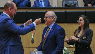Foto do conselheiro do TCE/SC Wilson Wan-Dall recebe do presidente da Alesc, deputado Mauro de Nadal, a medalha de homenagem à participação na Comstituição Estadual. São os dois homens brancos, que vestem terno azul. Ao lado, uma mulher de cabelos compridos e castanhos auxilia.