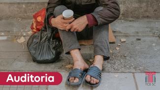 Foto de uma pessoa em situação de rua sentada sobre uma calçada. A foto está cortada, de modo que só é possível ver seus braços e pernas. Ela segura um copo descartável e sacos pretos. Sobre a imagem, no canto inferior esquerdo, o texto “Auditoria”, em tarja vermelha. No canto inferior direito, logo do TCE/SC. 