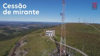 Imagem uma construção de dois andares ao centro. À direita dessa construção há uma torre. Ao fundo, também à direita, há mais algumas torres de transmissão. O cenário mostra o horizonte, com o céu azul e vegetação verde. Sobre o céu azul, escrito em branco, há à esquerda a expressão “Cessão de Mirante”. À direita, também sobre o céu azul, está o logotipo do TCE/SC