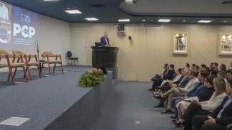 Banner horizontal com foto de um auditório durante um evento institucional. Em destaque, o presidente Herneus está em pé, atrás de um púlpito sobre o palco, enquanto o público acompanha a apresentação sentado em fileiras à direita da imagem. No telão ao fundo aparecem a identidade visual do evento e a sigla “PCP”. O palco conta com poltronas para debatedores