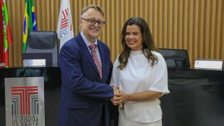 Foto do procurador-corregedor Diogo Ringenberg e da procuradora-geral Cibelly Farias durante o ato de posse. Eles estão em pé, um ao lado do outro, em frente ao plenário do TCE/SC. Estão sorrindo e se cumprimentam com um aperto de mãos. 
