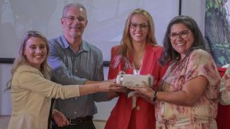 Imagem mostra quatro pessoas sorrindo. Da esquerda para direita, uma mulher de casaco bege, um homem de camisa cinza, uma mulher de casaco vermelho e uma mulher com roupa estampada