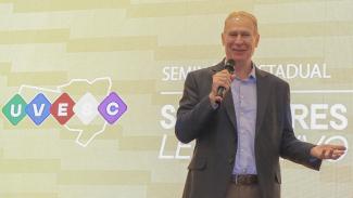 Imagem mostra um homem falando ao microfone. Ele veste um casaco cinza. Ao fundo, há um painel de luzes