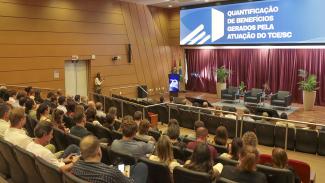 Banner horizontal com foto dos participantes do evento de treinamento no auditório do TCE/SC, entre diretores, servidores e autoridades do Tribunal, acomodados em poltronas enquanto acompanham a apresentação no palco. Eles estão de costas. Ao fundo, o púlpito, as bandeiras institucionais e o painel com o tema “Quantificação de benefícios gerados pela atuação do TCE/SC”, em ambiente formal e organizado.