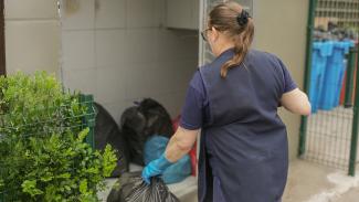 Foto de uma colaboradora, de costas, carregando sacos de lixo. Ela veste uniforme azul e usa luvas. 