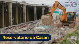 Imagem mostra o local do acidente, com uma estrutura de paredes caídas à esquerda e uma retroescavadeira amarela à direita. No canto inferior esquerta está escrito Reservatório da Casan em letras brancas sobre fundo preto.