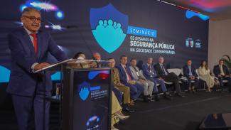 Imagem mostra em primeiro plano o conselheiro Aderson Flores, à esquerda. Ele está de óculos, veste um paletó cinza e fala ao microfone, que está em um púlpito. Ao fundo, na parede, um painel escuro com informações sobre o seminário. Também ao fundo, há autoridades, que estão sentadas