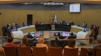 Imagem mostra o plenário do TCE/SC. As paredes são amareladas. Em frente, há uma mesa em forma de U onde ficam os conselheiros. Há televisões nas paredes onde ficam projetadas as imagens da própria sessão.