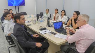 Foto mostra 10 pessoas em volta de uma mesa, durante reunião. Sobre a mesa de madeira clara há um laptop e alguns blocos de anotação. À direita, de frente para câmara, há quatro mulheres. Na cabeceira há um homem de camisa rosa 