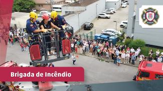 Banner com a foto da simulação. Em primeiro plano, dois bombeiros militares e uma “vítima” sendo resgatada pela escada do Corpo de Bombeiros. Ao fundo, servidores do TCE/SC, na calçada, assistem à operação. Sobre a foto, tarja bordô com o texto Brigada de Incêndio. E no canto superior direito, o logo da Brigada de Incêndio. 