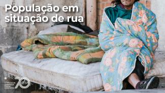 Imagem mostra uma pessoa à direita, enrolada em um cobertor. Ao lado da pessoa há um colchão com outro cobertor sobre ele. Ao fundo aparece um muro de uma área externa