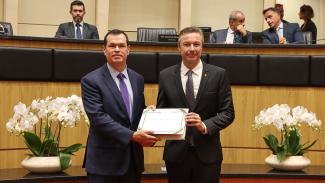 Banner horizontal com foto do conselheiro Adircélio de Moraes ao lado do deputado Napoleão Bernardes durante a sessão na Alesc. Eles estão em pé, segurando, juntos, o título concedido ao conselheiro. Vestem terno e gravata e sorriem para a foto. Ao fundo, a bancada com demais autoridades presentes na sessão.