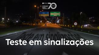 Imagem feita á noite mostra uma via iluminada, com as luzes dos faróis iluminando as placas de indicação de trânsito. Ao centro, escrito em branco, está escrito “teste em sinalizações”. No alto, ao centro, está o logo de 70 anos do TCE/SC