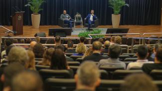 Banner horizontal com foto, em primeiro plano, da plateia, de costas, sentada em poltronas no auditório do TCE/SC. À frente do público, no palco, estão os palestrantes: o advogado Bruno Peixoto e o engenheiro eletricista Willian Pinheiro. Ambos estão sentados em poltronas pretas e falam ao público.  