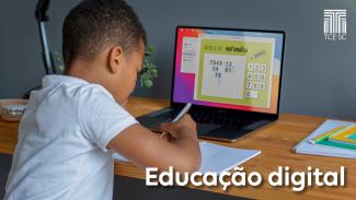 Imagem mostra uma criança de costas, vestindo camiseta branca e em frente a um computador que está aberto sobre uma mesa. A criança segura um lápis e escreve em um caderno. O computador mostra uma conta de matemática em sua tela.