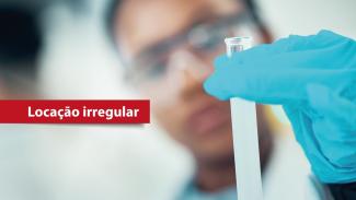 Banner horizontal que mostra, em primeiro plano, uma mão com luva azul segurando um tubo de ensaio com líquido claro. Ao fundo, desfocado, aparece uma pessoa usando óculos de proteção em ambiente que lembra um laboratório. À esquerda da imagem há uma faixa vermelha com o texto “Locação irregular”. 