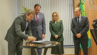 Foto do vice-presidente do TCE/SC, conselheiro José Nei Ascari, em pé, assinando o termo, ao lado de outras três autoridades: desembargadores Francisco Oliveira Neto e Amarildo Carlos de Lima e desembargadora Maria do Rocio. Ascari é um homem branco, de cabelos grisalhos. Veste terno e gravata e usa óculos de grau