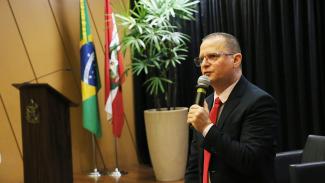 Foto do auditor fiscal de controle externo Alcionei Vargas de Aguiar. Ele é um homem branco, de cabelos escuros e raspados. Ele usa terno e gravata e óculos de grau. Está em pé, no palco do auditório do TCE/SC, durante o evento. Segura o microfone com uma das mãos, enquanto fala.